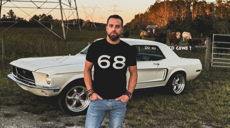 David in a black '68' shirt in front of the white Mustang at sunset